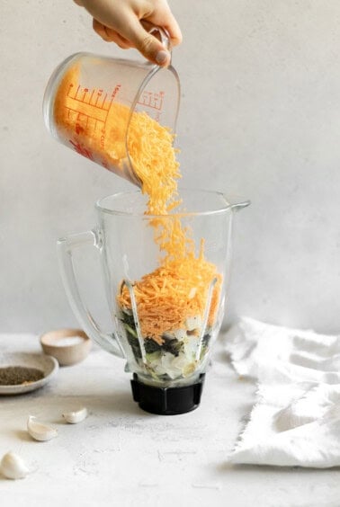 A person adding shredded cheese to a blender for broccoli cheese soup.