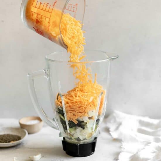 A person adding shredded cheese to a blender for broccoli cheese soup.