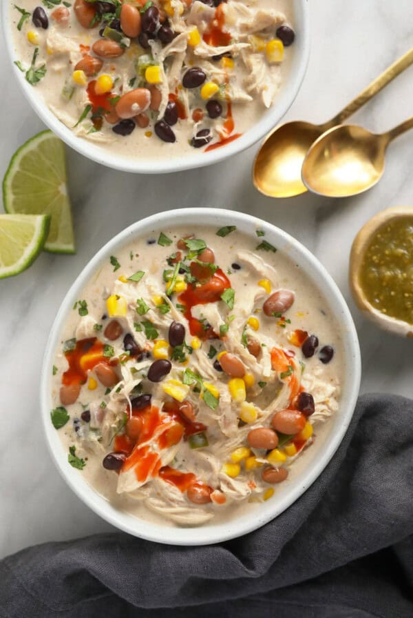 Two bowls of Mexican chicken soup, garnished with cream cheese and chili, placed on a marble table.