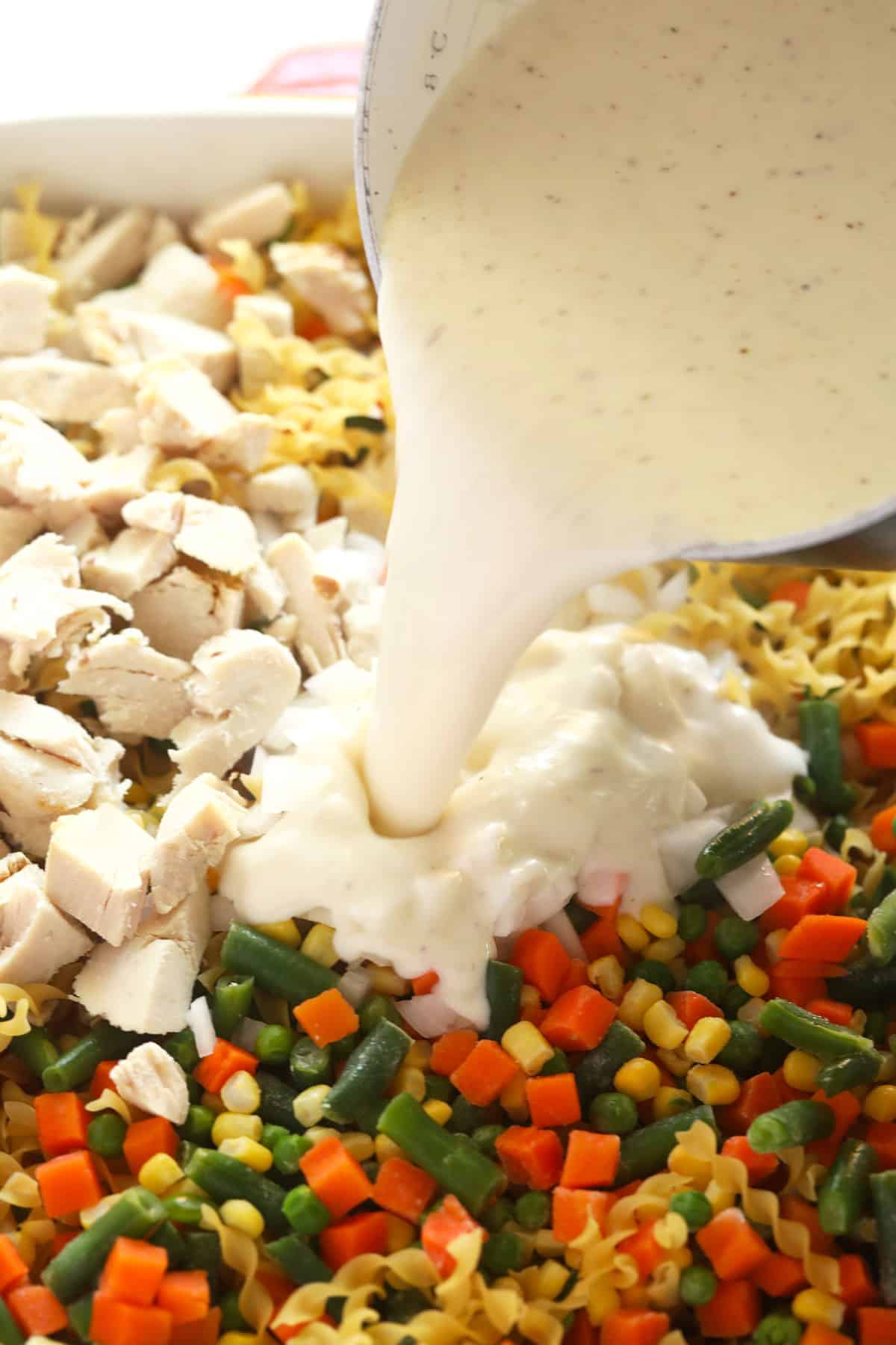 Creamy cheese sauce being poured over a chicken noodle casserole before being baked.