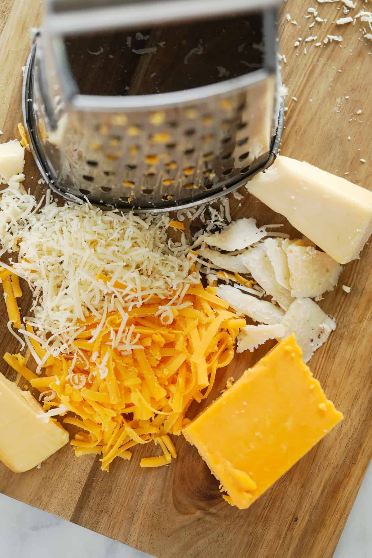 A box grater with different kinds of shredded cheese on the cutting board. 