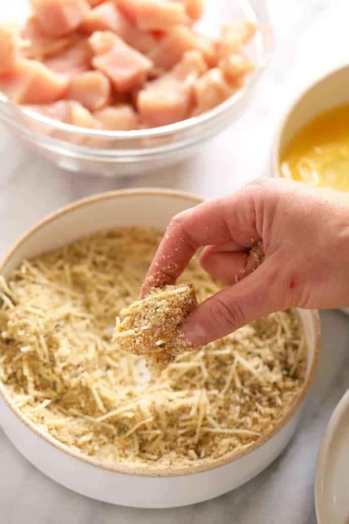 A hand dipping a piece of chicken into bread crumbs. 