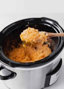 A wooden spoon stirs a crockpot of cheesy macaroni and cheese.