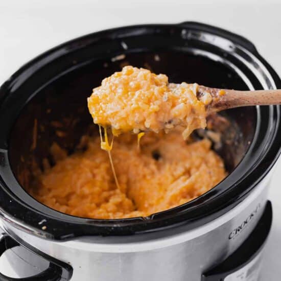 A wooden spoon stirs a crockpot of cheesy macaroni and cheese.