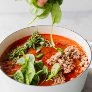 A person preparing Lasagna Soup by squeezing fresh spinach into a pot of tomato sauce.