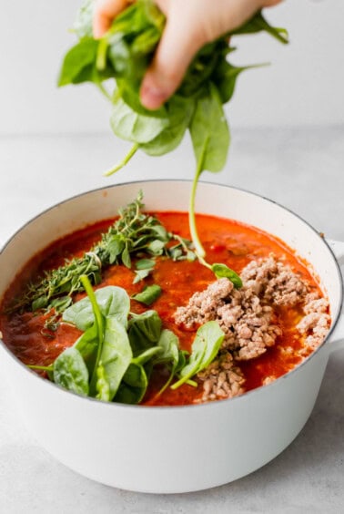 A person preparing Lasagna Soup by squeezing fresh spinach into a pot of tomato sauce.