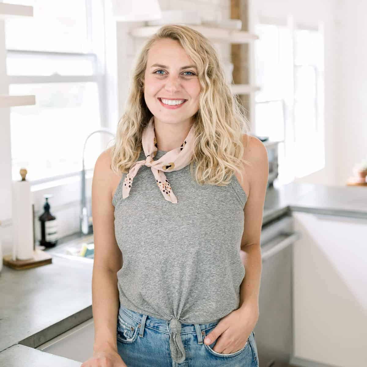 a blonde woman in a gray tank top and jeans standing in a kitchen, passionate about cheese.