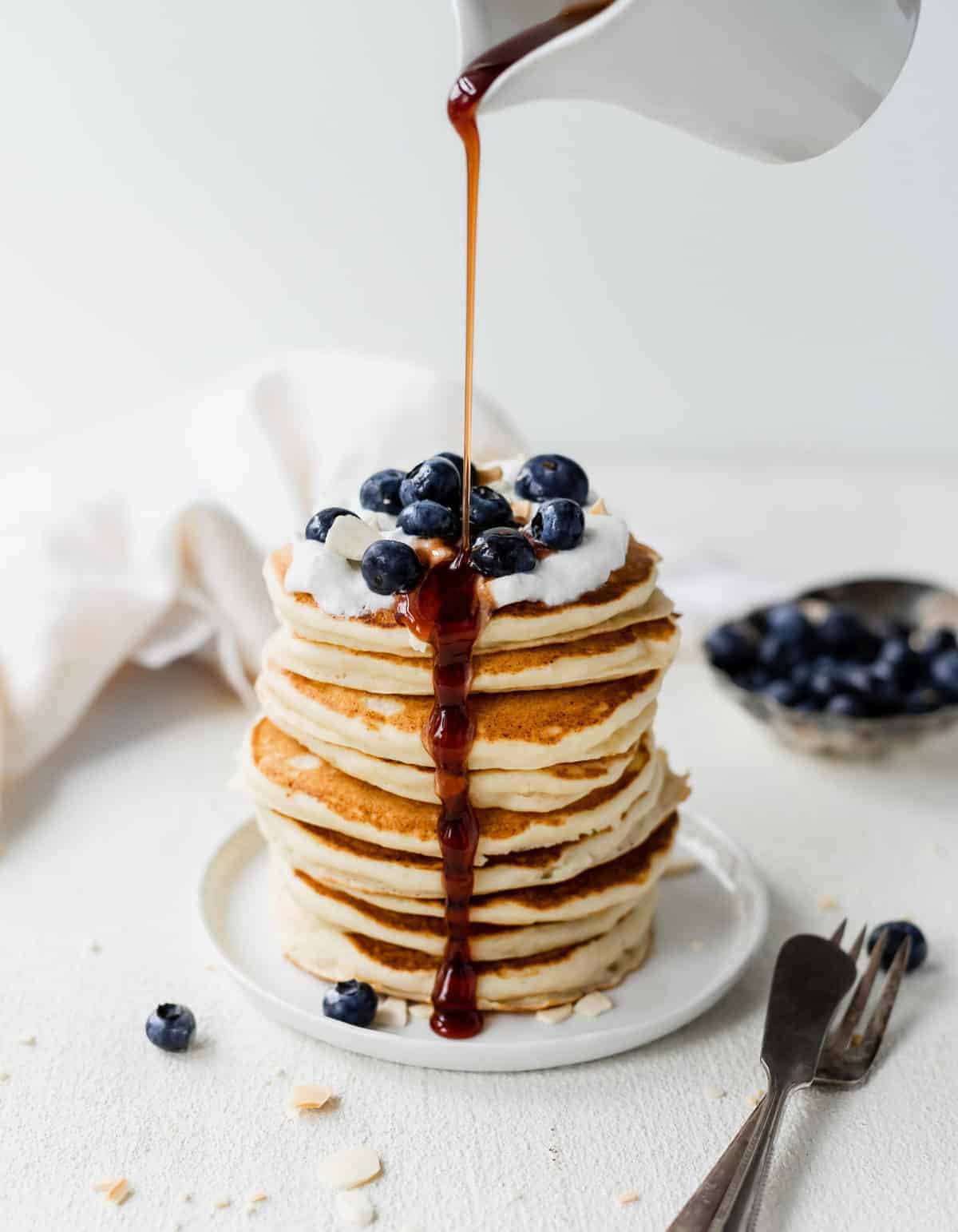 Maple syrup being poured over a stack of ricotta pancakes on a plate.