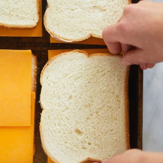 a person cutting a piece of cheese and placing it between two slices of bread on a sheet pan to make grilled cheese.