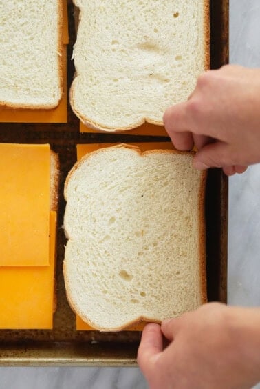 a person cutting a piece of cheese and placing it between two slices of bread on a sheet pan to make grilled cheese.