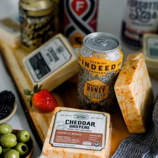 blocks of cheese and cans of beer on a wooden cutting board surrounded by olives and fruit