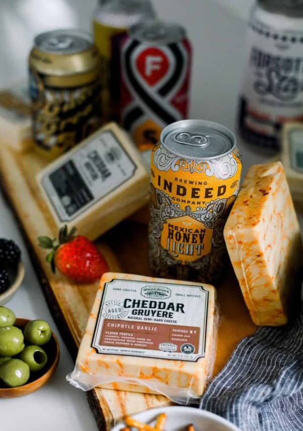 blocks of cheese and cans of beer on a wooden cutting board surrounded by olives and fruit