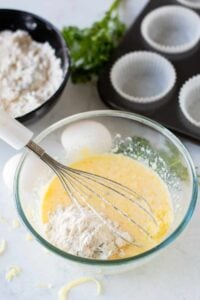 A bowl with eggs, flour, and a whisk used to make cheese muffins.