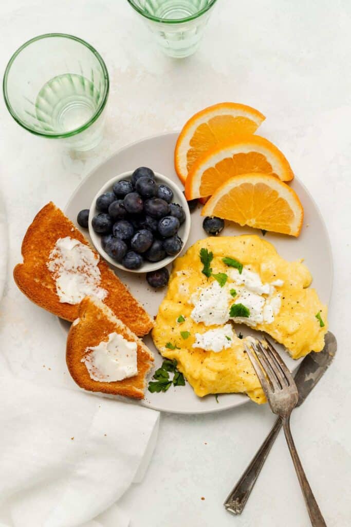 Goat cheese scrambled eggs on a plate with toast, blueberries, and oranges.