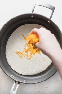 a person putting cheese on a tortilla in a pan.