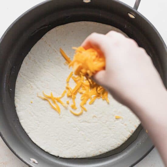 a person putting cheese on a tortilla in a pan.