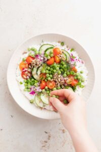 A person assembling a cottage cheese salad with vegetables and peas.
