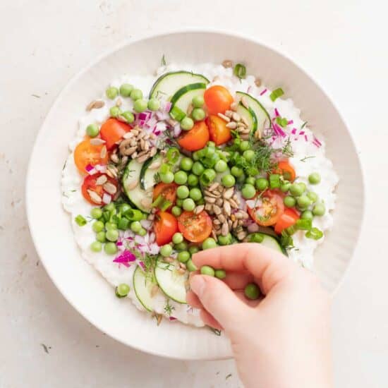 A person assembling a cottage cheese salad with vegetables and peas.