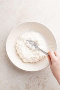 a person's hand is holding a spoonful of cottage cheese salad in a white bowl.