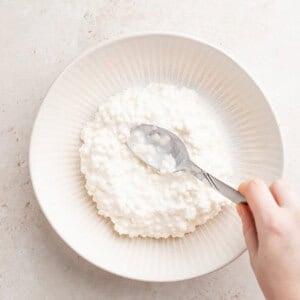 a person's hand is holding a spoonful of cottage cheese salad in a white bowl.