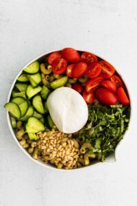 A pesto pasta salad with burrata served in a white bowl with tomatoes, cucumbers, and mozzarella.