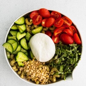 A pesto pasta salad with burrata served in a white bowl with tomatoes, cucumbers, and mozzarella.