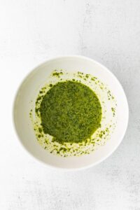 A bowl of green pesto with burrata on a white surface.