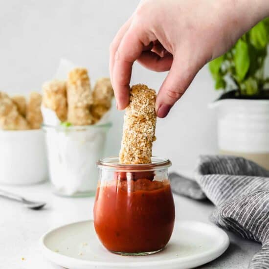 A person preparing homemade mozzarella sticks by dipping a stick into a jar of sauce.