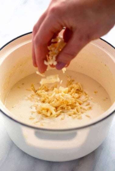 A person adding shredded cheese to a bowl of soup, reminiscent of baked mac and cheese.