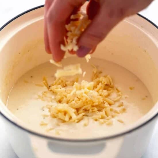 A person adding shredded cheese to a bowl of soup, reminiscent of baked mac and cheese.