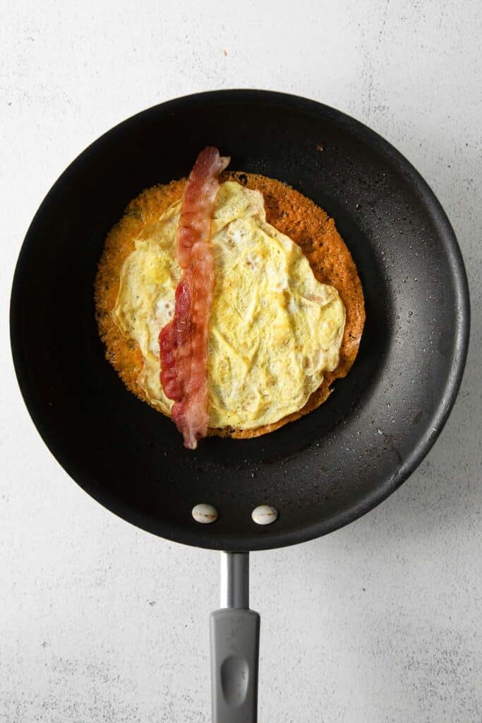 cheese wrap and egg topped with a strip of bacon in a skillet