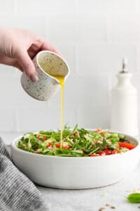 a hand is pouring olive oil over a salad in a white bowl.