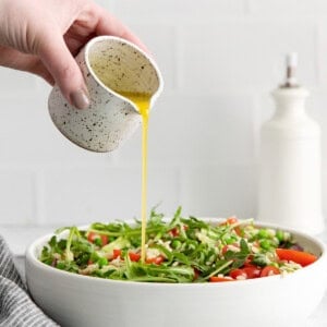 a hand is pouring olive oil over a salad in a white bowl.