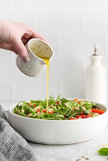 a hand is pouring olive oil over a salad in a white bowl.