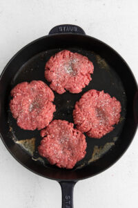 four hamburger patties in a skillet on a white surface.