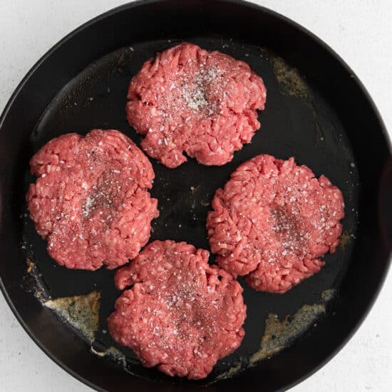 four hamburger patties in a skillet on a white surface.