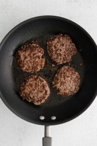 four hamburger patties in a frying pan on a white background.