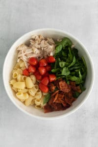 a white bowl filled with pasta, chicken, spinach and tomatoes.