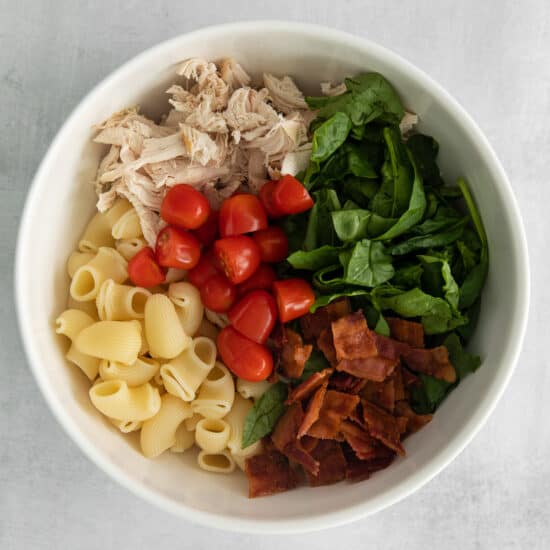 a white bowl filled with pasta, chicken, spinach and tomatoes.