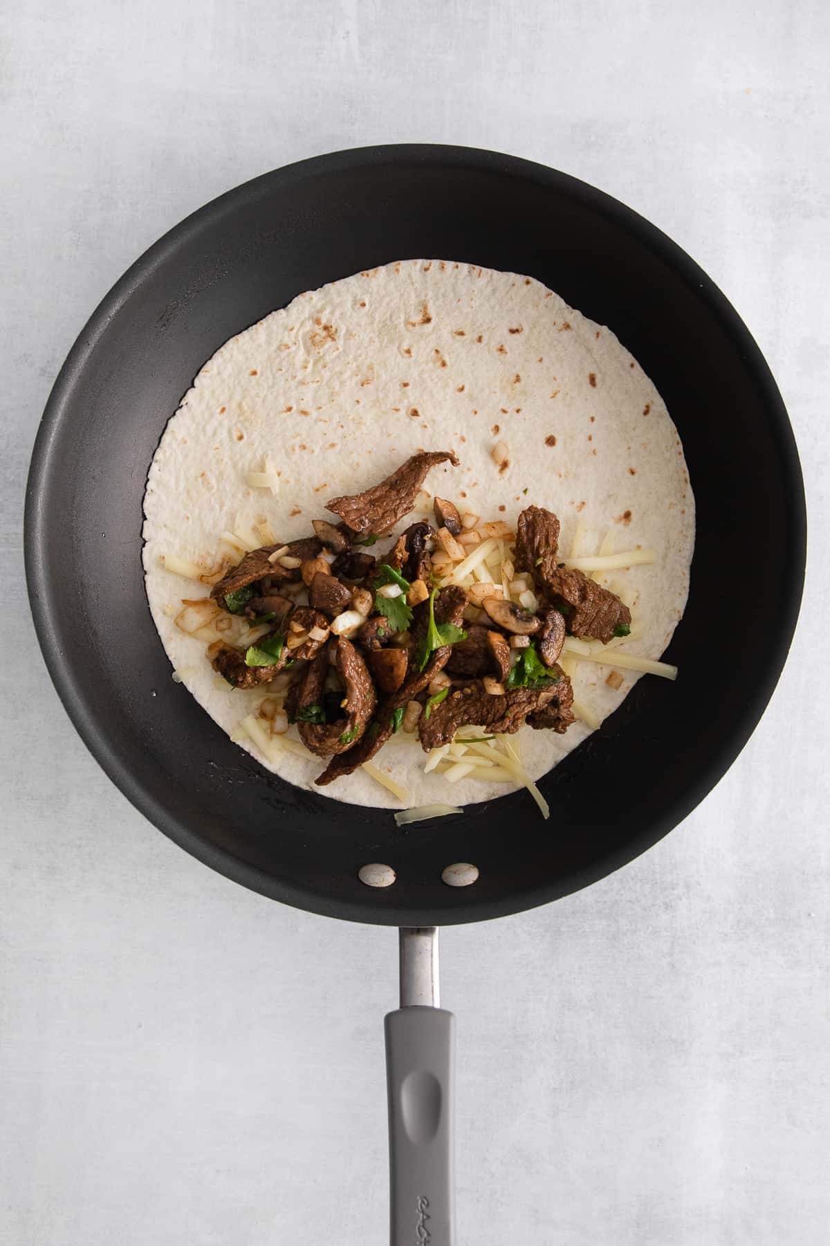 Cheese, steak, and onions on a tortilla in a skillet.