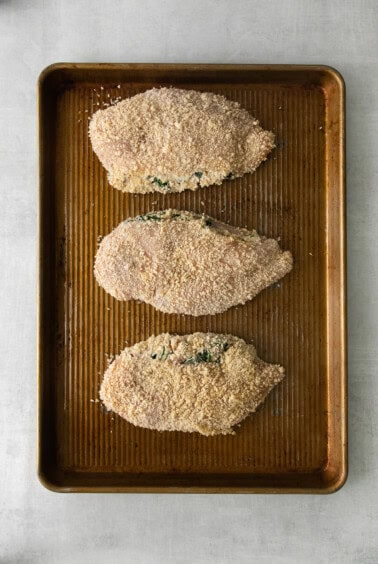 three breaded chicken breasts on a baking sheet.