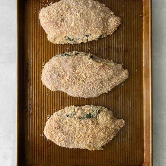 three breaded chicken breasts on a baking sheet.