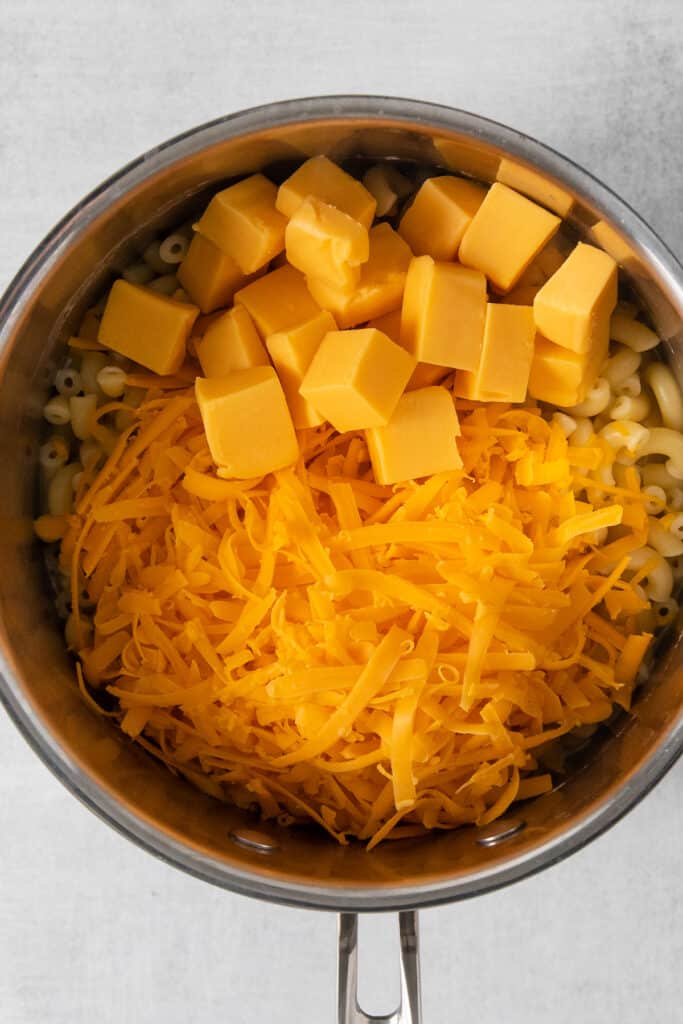 Shredded and cubed cheese in a pot with macaroni noodles.