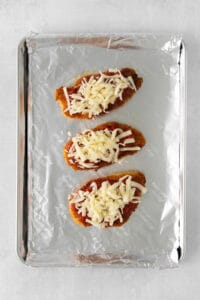 three chicken breasts topped with cheese on a baking sheet.