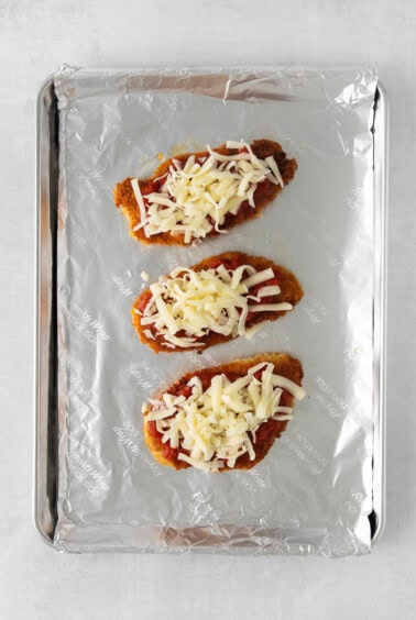 three chicken breasts topped with cheese on a baking sheet.