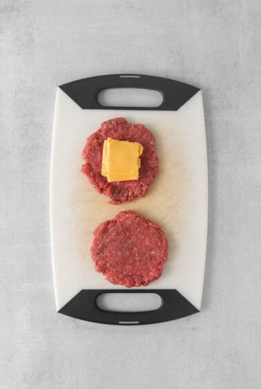 two hamburger patties on a cutting board with cheese.
