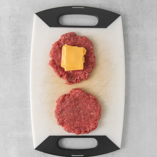 two hamburger patties on a cutting board with cheese.
