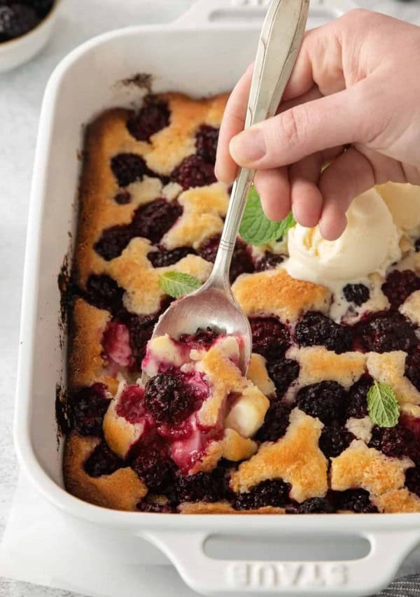 a person scooping blackberry cobbler from a baking dish.