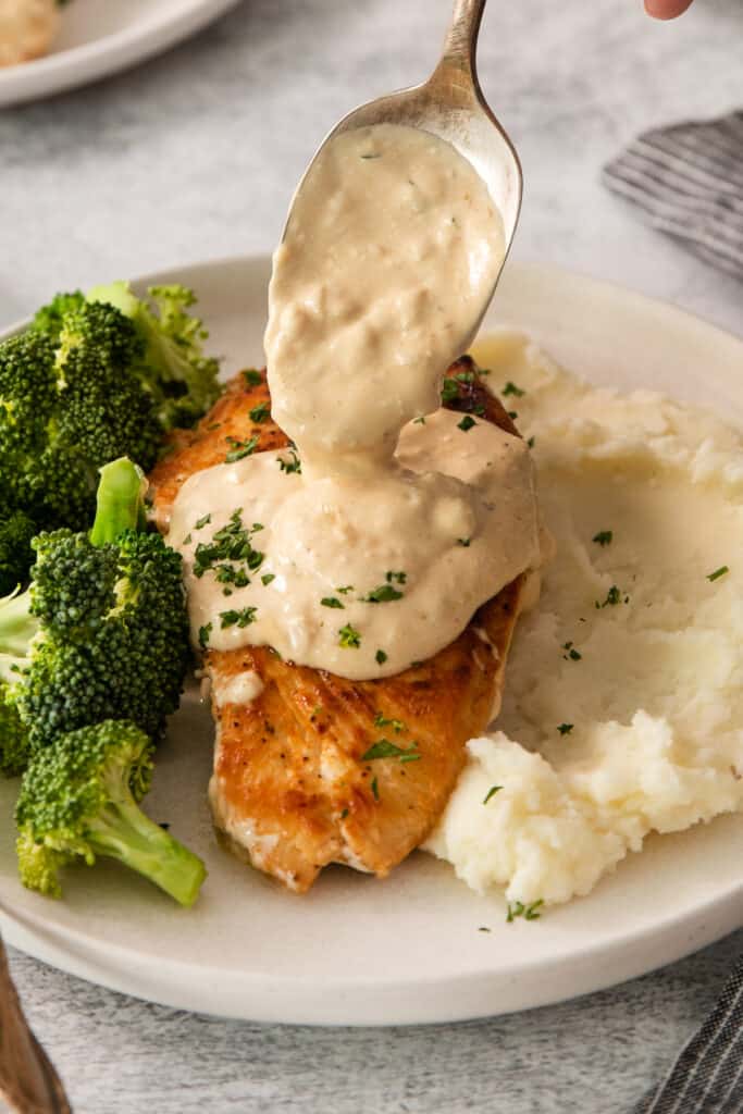 a person pouring a sauce over chicken and broccoli on a plate.