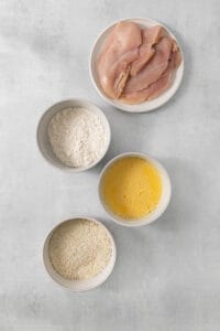 four bowls with chicken, flour, eggs and oil on a grey background.